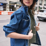 woman wearing a blue denim jacket with a frill collar, white stitch detailing, button-up front, and four large pockets with a green and white striped denim shirt and jeans, and a denim bucket hat on a new york city street
