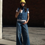 woman wearing a blue denim vest/ waistcoat with our signature white stitch detailing, two large patch pockets, and a frill collar with blue jeans against a brick background
