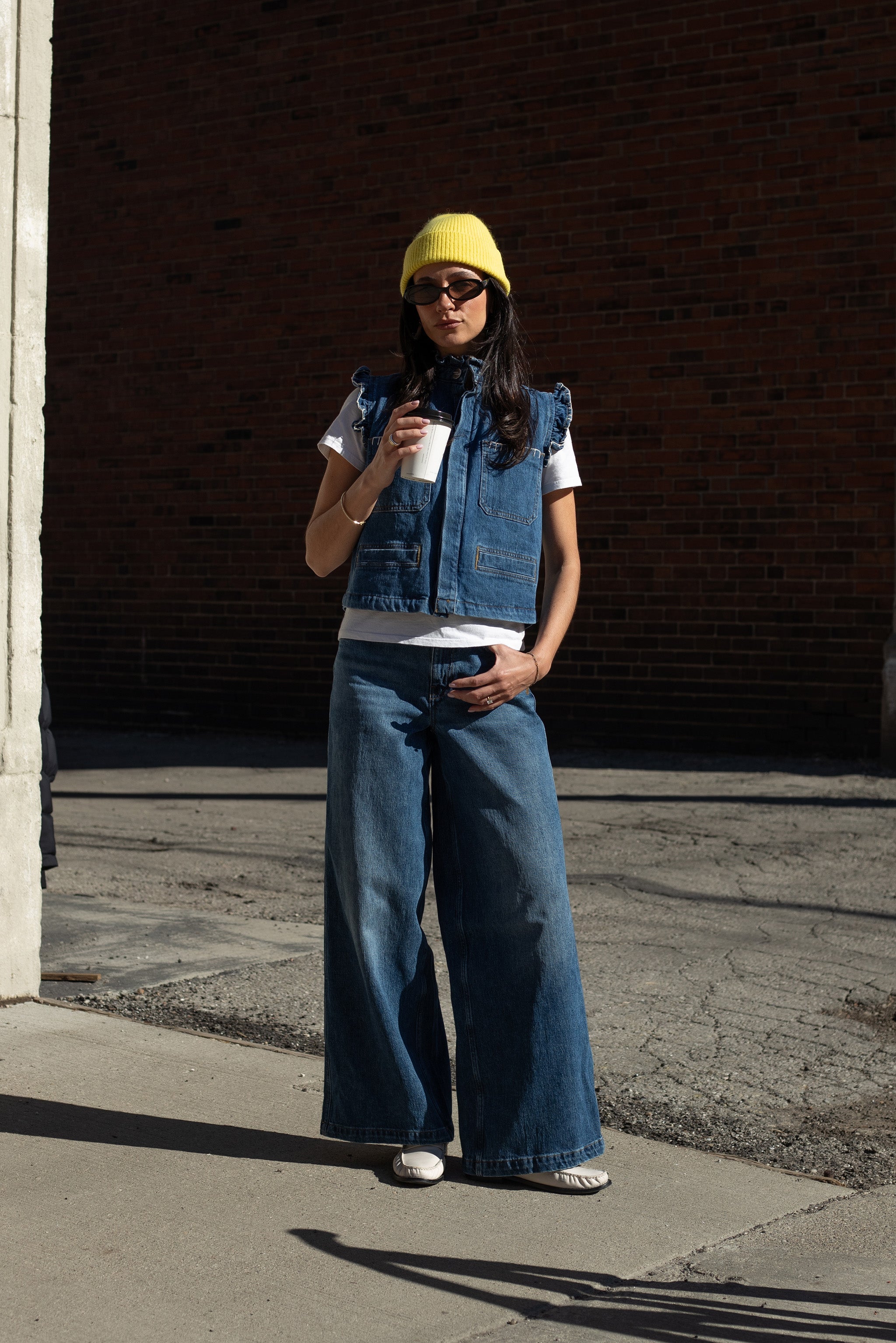 woman wearing a blue denim vest/ waistcoat with our signature white stitch detailing, two large patch pockets, and a frill collar with blue jeans against a brick background