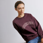 Model wearing bordeaux embroidered sweatshirt with statement logo and casual relaxed fit, styled with neutral bottoms.