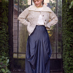 Woman wearing a textured sweater and blue skirt standing in front of a glass door with greenery around.
