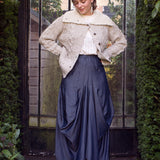 Woman wearing a textured sweater and blue skirt standing in front of a glass door with greenery around.