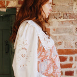 Seated pose featuring white blouse with long sleeves, embroidery detail, and scalloped trim edges.