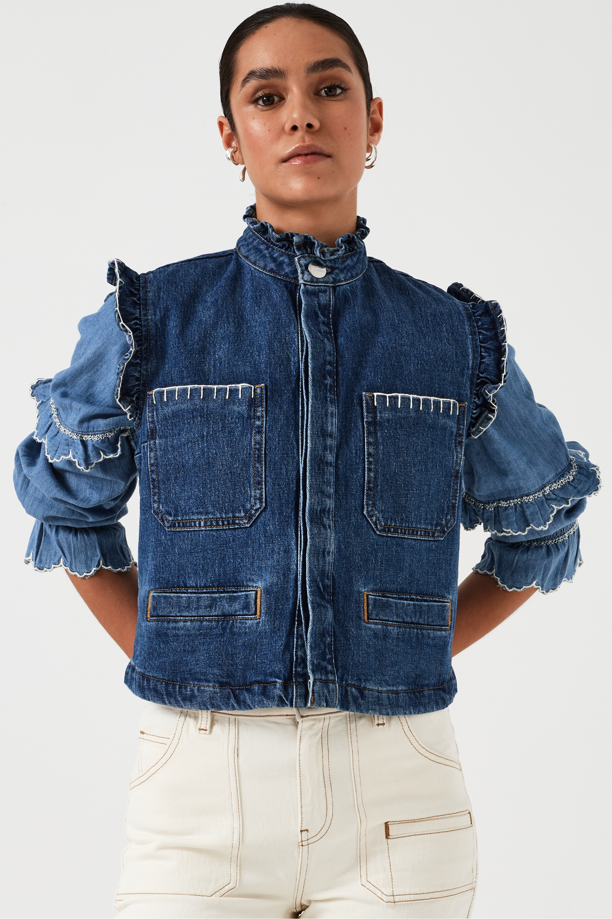 woman wearing a blue denim vest/ waistcoat with our signature white stitch detailing, two large patch pockets, and a frill collar with a blue shirt and white jeans against a white background