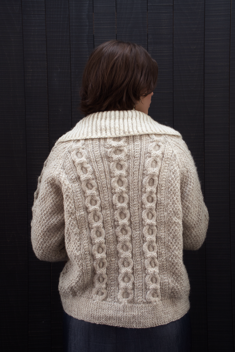 Person wearing a beige knitted cardigan with intricate patterns against a dark background