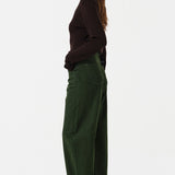 Woman wearing dark green barrel leg jeans and a brown shirt on a white background, photographed from the side