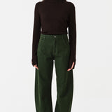 Woman wearing dark green barrel leg jeans and a brown shirt on a white background