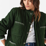 Close up of woman wearing a quilted dark green jacket with white embroidered details and blue jeans on a white background