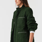 Woman wearing a quilted dark green jacket with white embroidered details and blue jeans on a white background