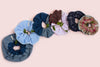 A selection of Saywood scrunchies laid together on a pink background, with green foliage winding through.